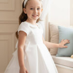 فستان بيبي بلون الكريمي مع تفاصيل فيونكة ورباط رأس للأعمار من 6 إلى 24 شهراً